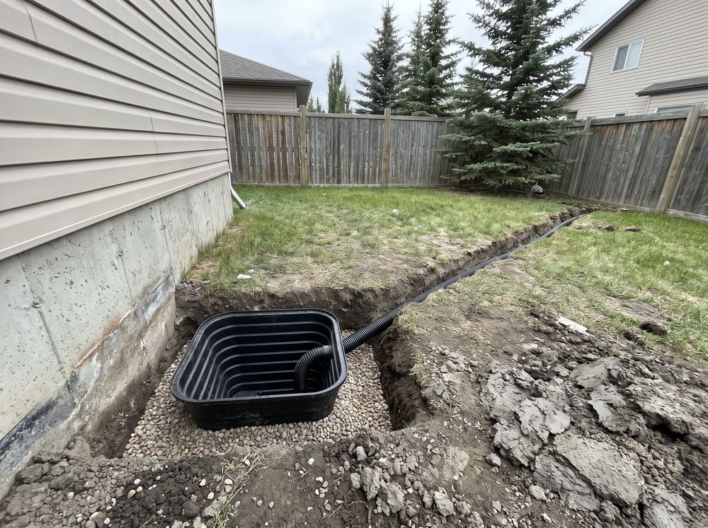 Square sump pit with a plastic basin on gravel beside a house foundation and a discharge line trench