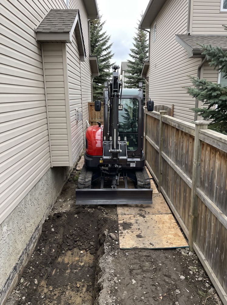 Excavator parked in the narrow gap between two houses with a trench dug ahead of it