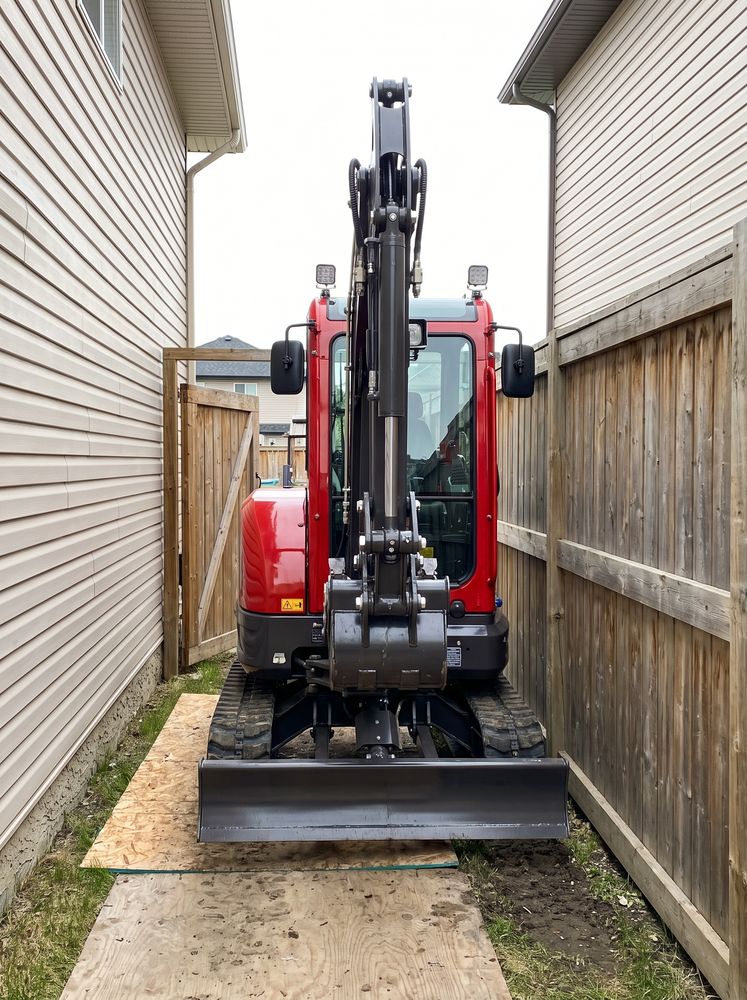 Compact excavator on plywood mats squeezing through a narrow side yard between a house and fence